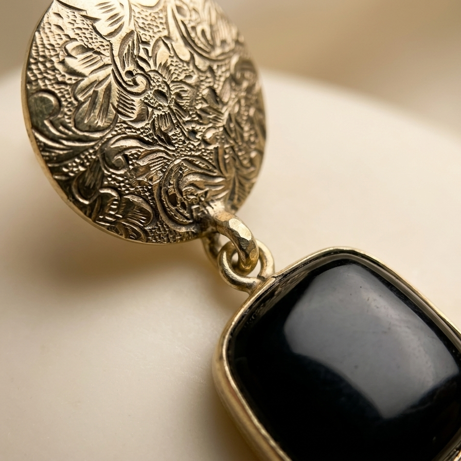 A close-up of an ornate gold-toned earring featuring a round, intricately engraved disc and a dangling square black stone, displayed on a soft cream-colored surface.