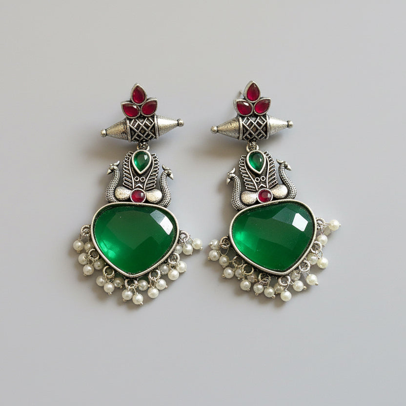 Pair of ornate earrings with green heart stones and red accents on a light gray background