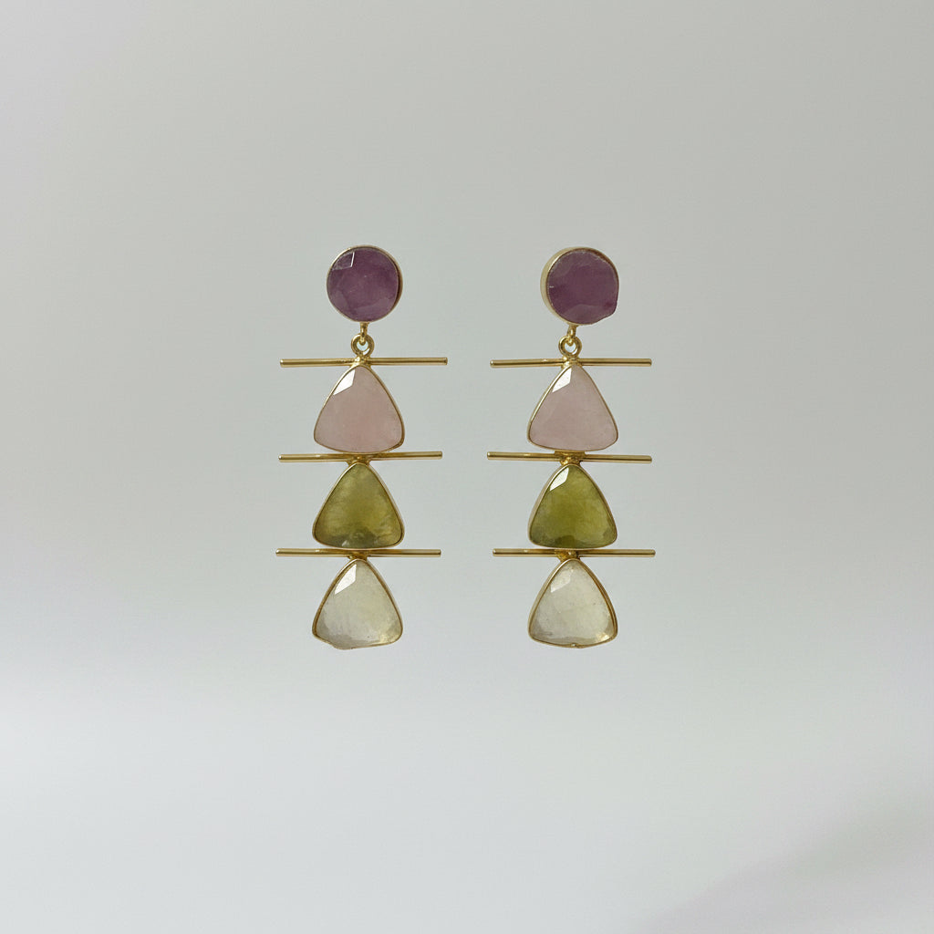A pair of gold drop earrings with stacked triangular gemstone charms in pale pink, yellow-green, and cream, topped with round dusty pink studs, displayed on a black velvet jewelry stand.