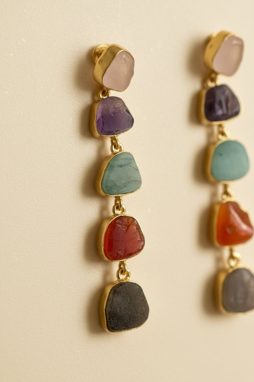 A pair of gold drop earrings featuring five small, irregular natural gemstones in purple, green, orange, gray, and beige, displayed on a black textured fabric background.