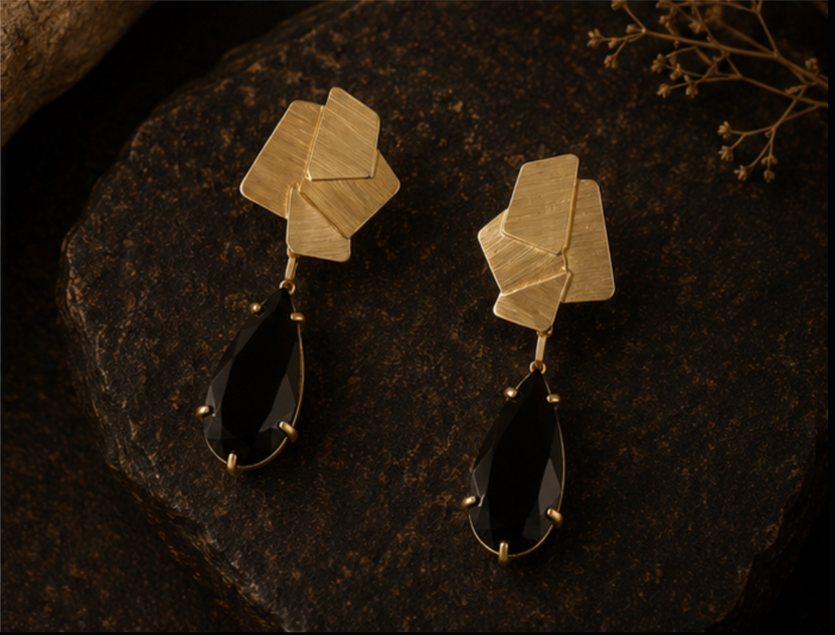 A pair of gold geometric drop earrings with layered hexagonal studs and teardrop-shaped black gemstones, displayed on a dark textured stone surface.