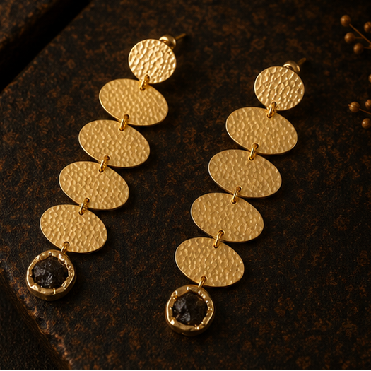 Gold-toned drop earrings with textured circular disc links and dark oval stones, shown on a display stand, worn by a model, and in a close-up detail shot against a dark brown background.