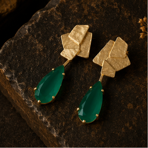 Geometric gold drop earrings with teardrop-shaped emerald green stones, shown on a gold display stand, worn by a model, and resting on a dark stone surface.