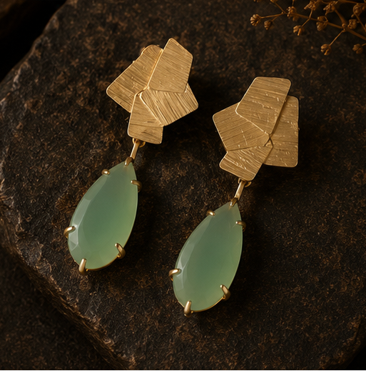 Geometric drop earrings with textured gold-tone metal and light green teardrop stones, shown on a display stand, worn by a model, and close-up on a dark stone surface.