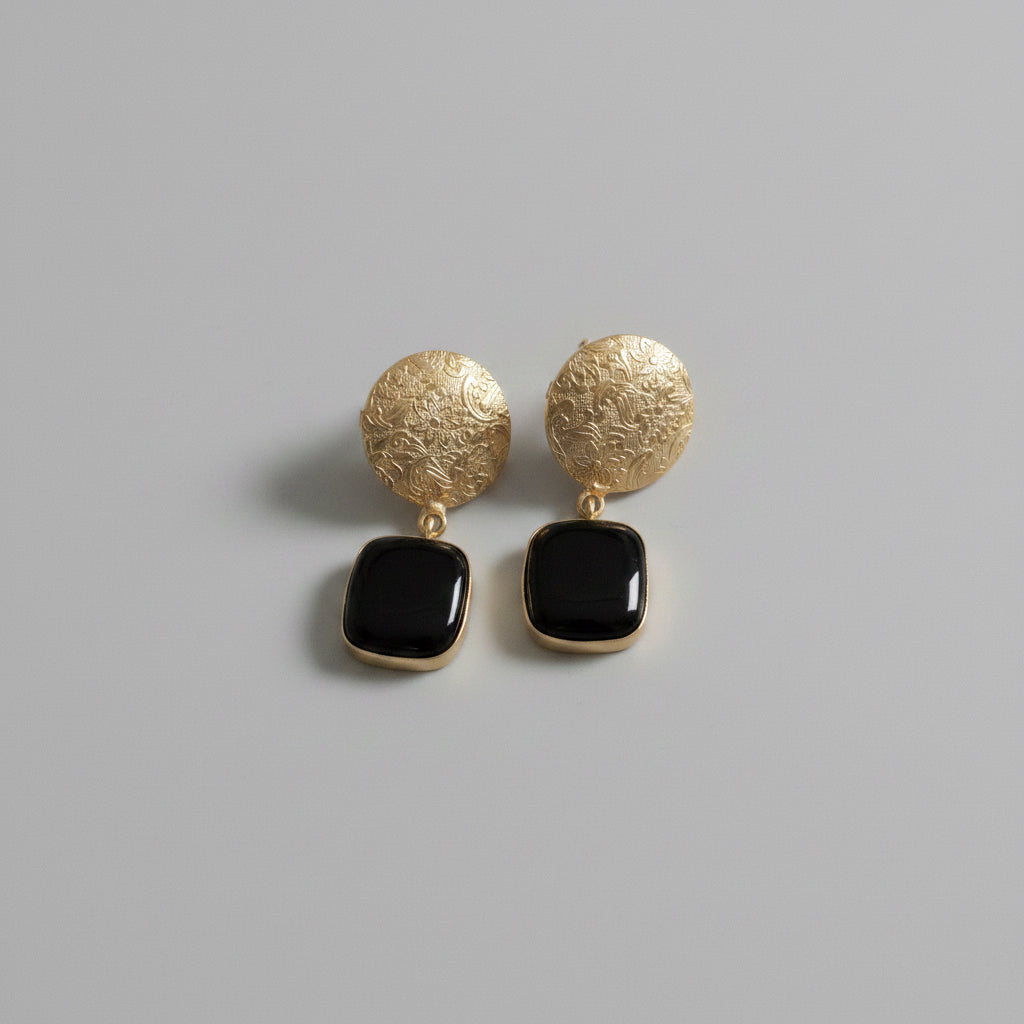 A pair of gold-toned drop earrings with textured circular studs and square black stones, displayed against a solid black background.