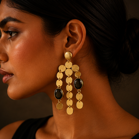 Gold-toned statement dangle earrings with circular coin-like discs and dark oval stones, shown on a display stand, worn by a model, and laid flat on a dark surface.