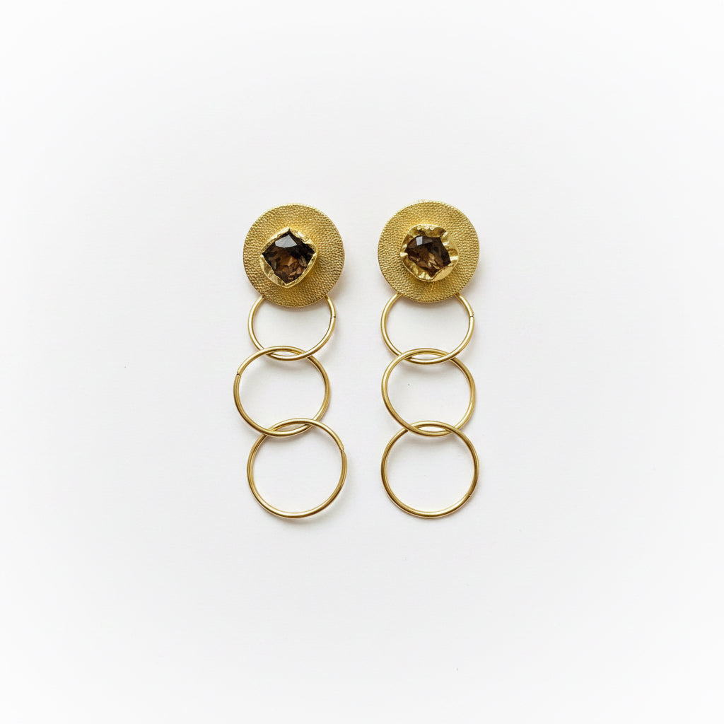 A pair of gold-toned earrings with circular textured studs featuring dark brown faceted gemstones and three linked oval rings, displayed on a black background.