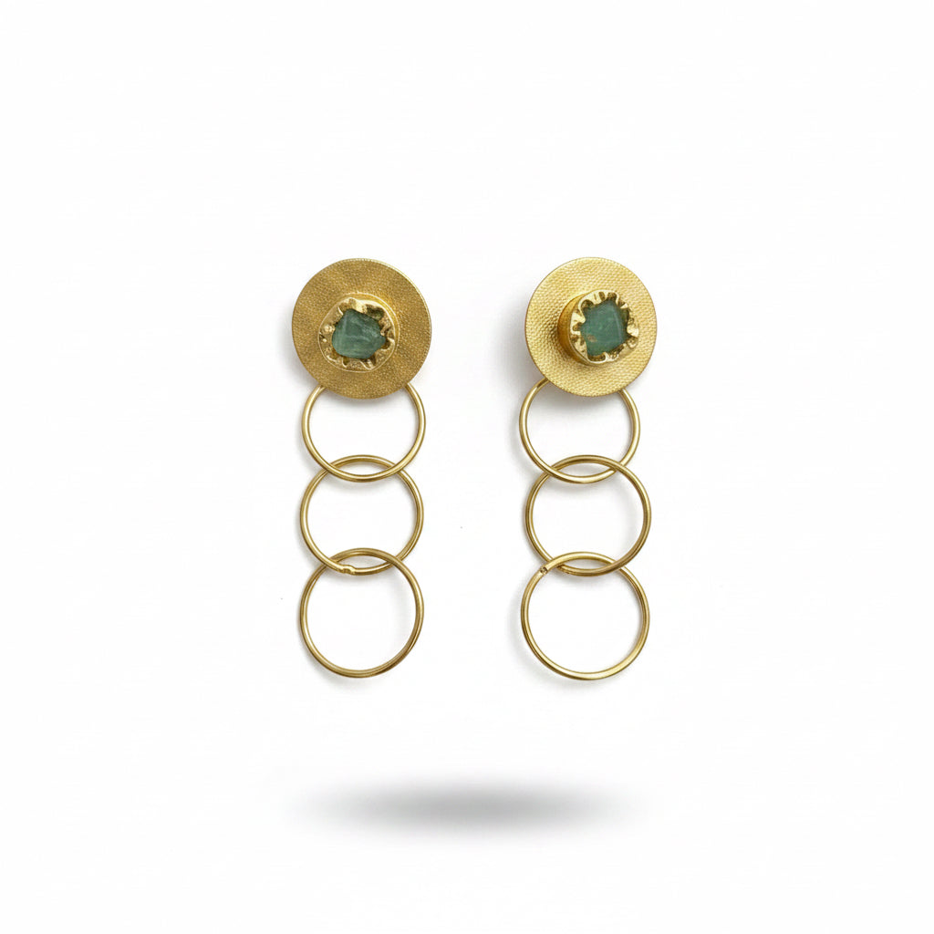 A pair of gold-toned drop earrings with textured circular studs, small green oval inlays, and three stacked hanging rings, displayed on a dark textured background.