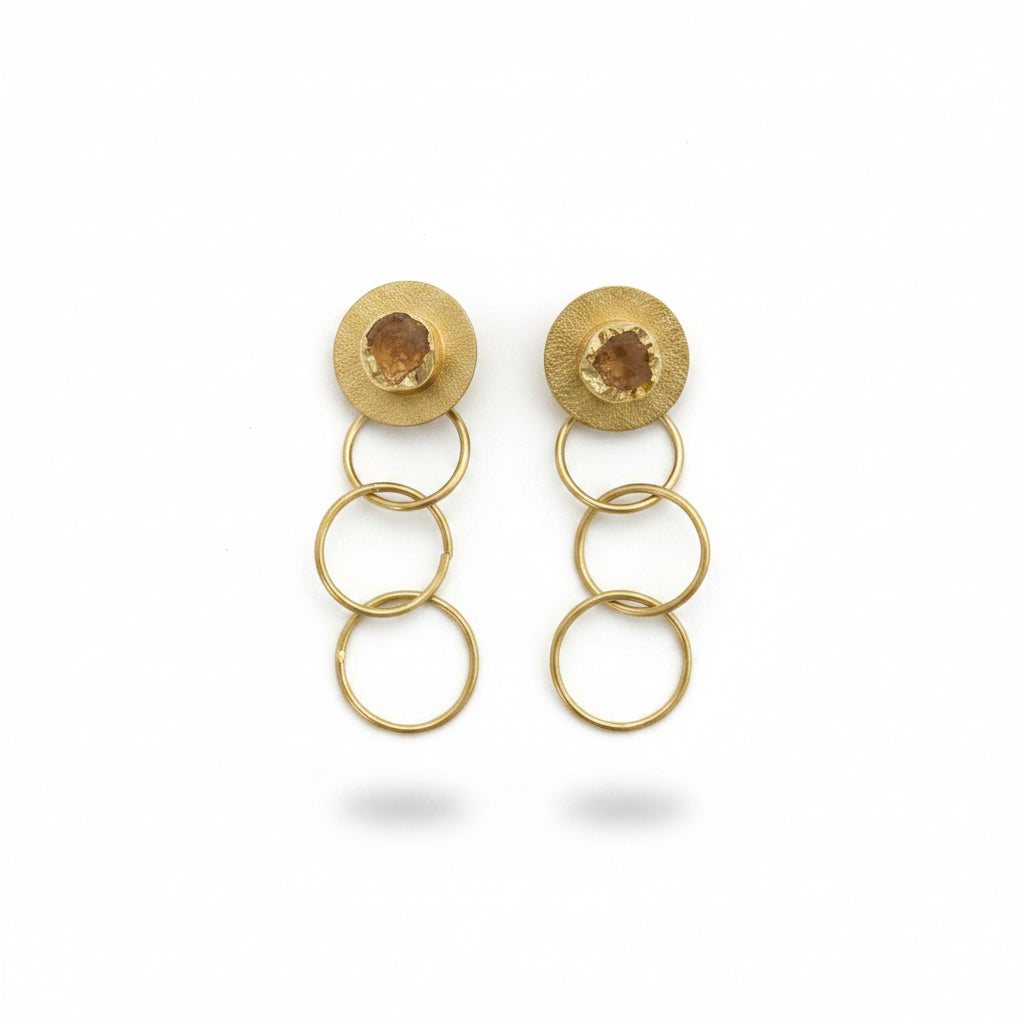 A pair of gold drop earrings with circular stacked rings and a small faceted stone on each post, displayed on a black textured background.