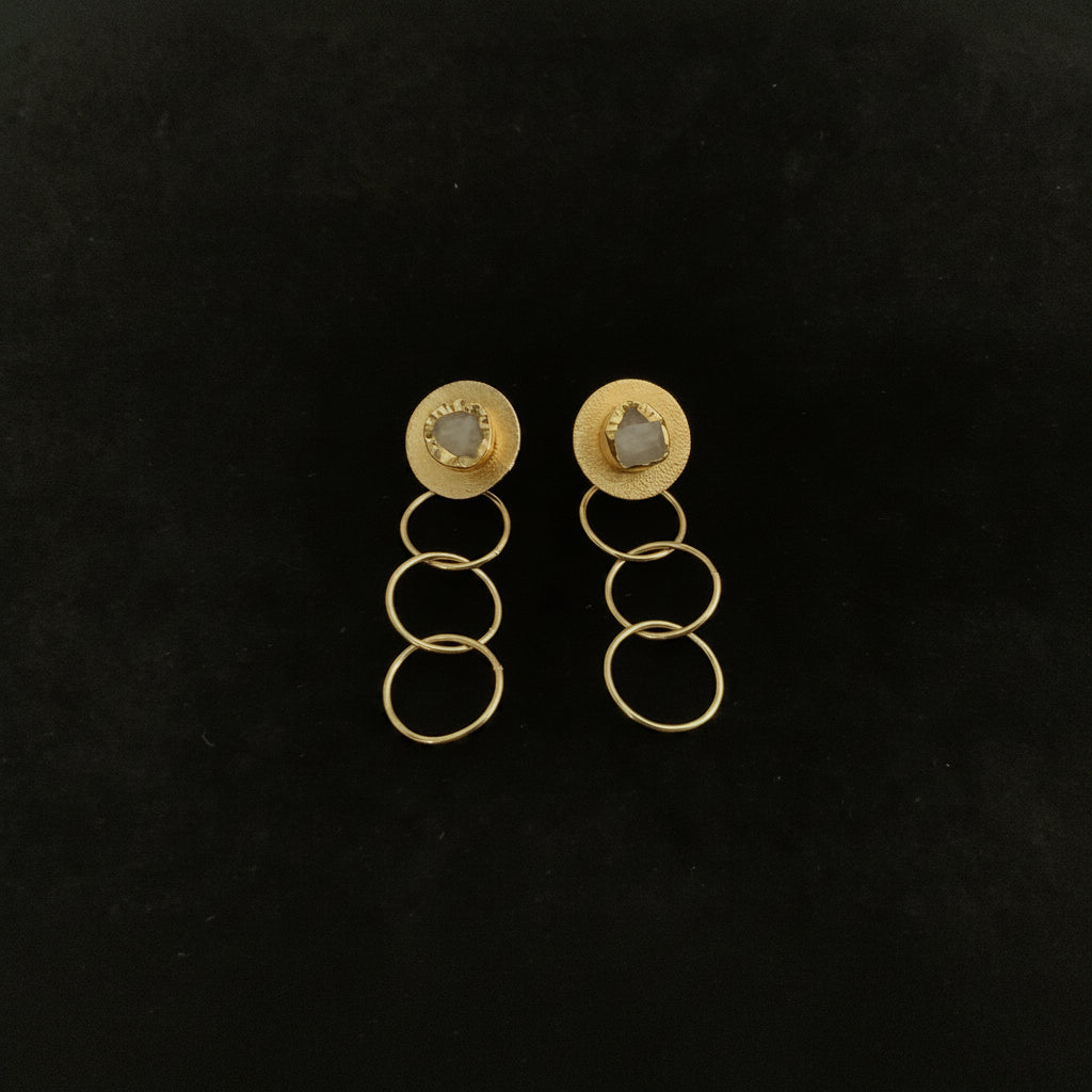A pair of gold-toned drop earrings with circular studs and three stacked hanging rings, displayed on a dark textured background.
