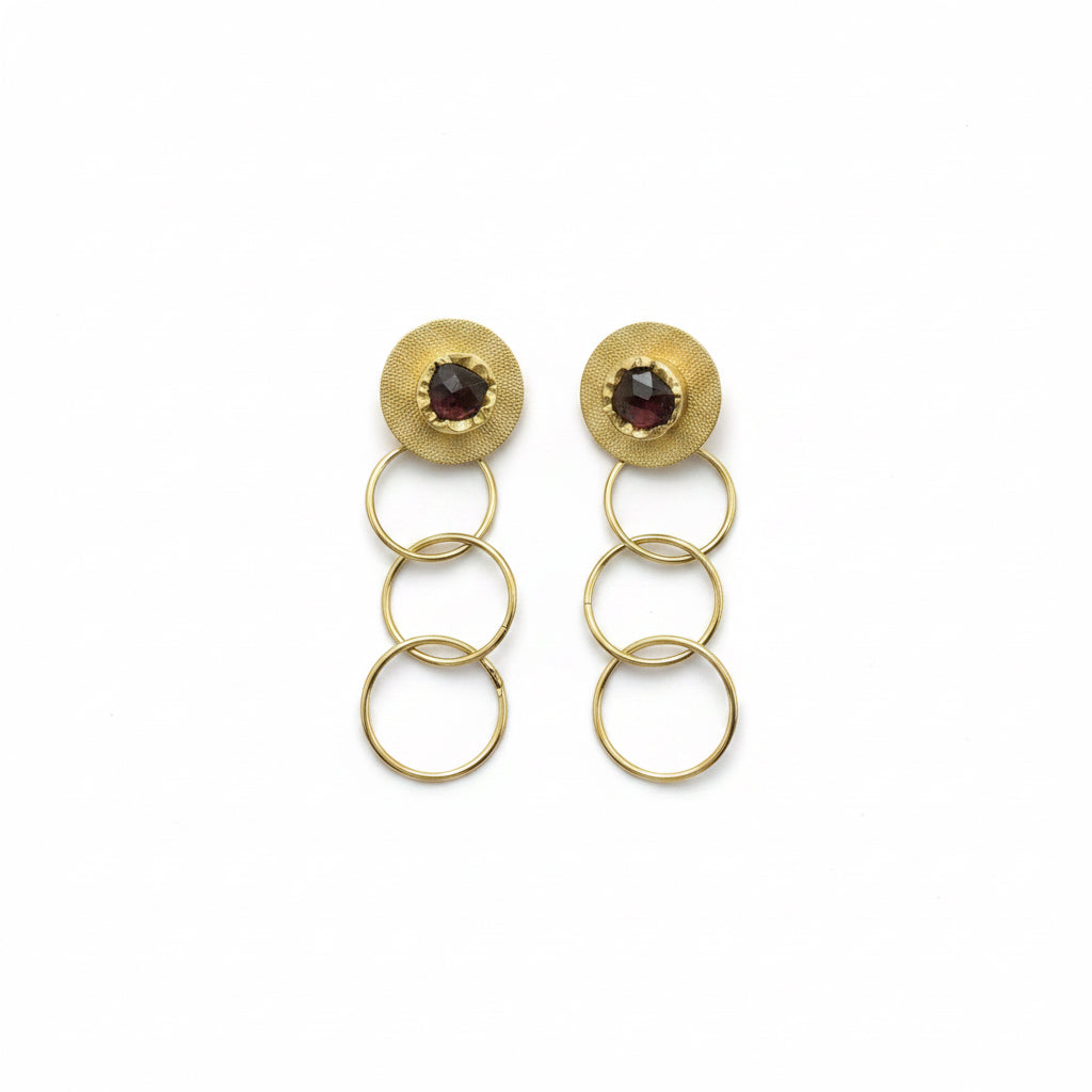 A pair of gold-toned dangle earrings with circular disc tops featuring small dark gemstones, suspended by three interlocking rings on a black background.