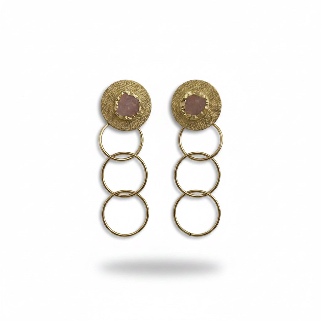 A pair of gold dangle earrings with circular disc tops featuring oval pinkish-purple gemstones, suspended by three interlinked rings on a black background.