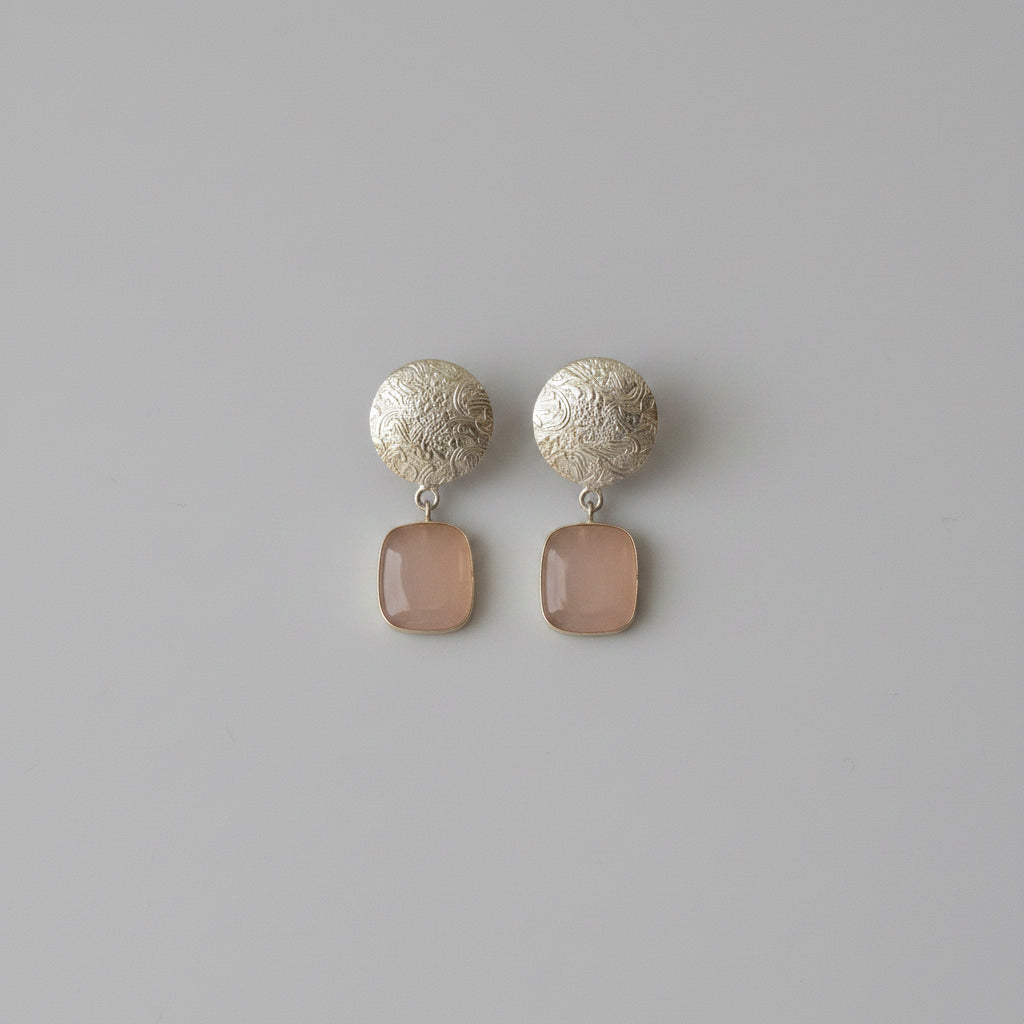 A pair of dangle earrings featuring textured silver circular studs and smooth, rectangular taupe-brown pendants, displayed against a solid black background.