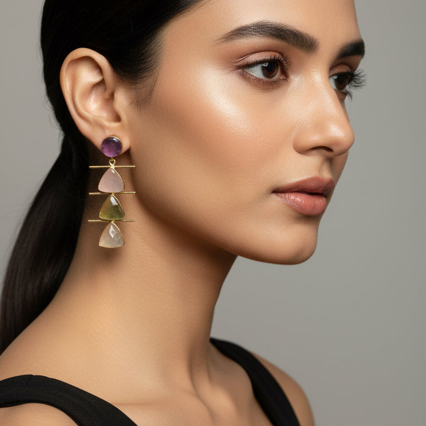 A pair of gold drop earrings with stacked triangular gemstone charms in pale pink, yellow-green, and cream, topped with round dusty pink studs, displayed on a black velvet jewelry stand.