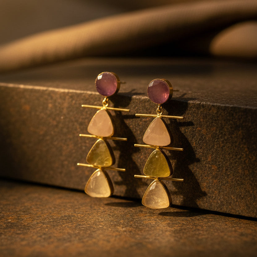 A pair of gold drop earrings with stacked triangular gemstone charms in pale pink, yellow-green, and cream, topped with round dusty pink studs, displayed on a black velvet jewelry stand.