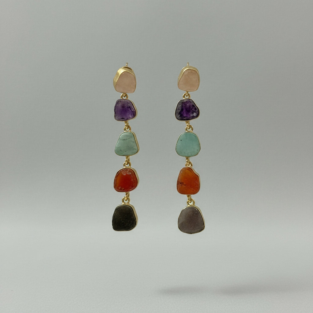 A pair of gold drop earrings featuring five small, irregular natural gemstones in purple, green, orange, gray, and beige, displayed on a black textured fabric background.