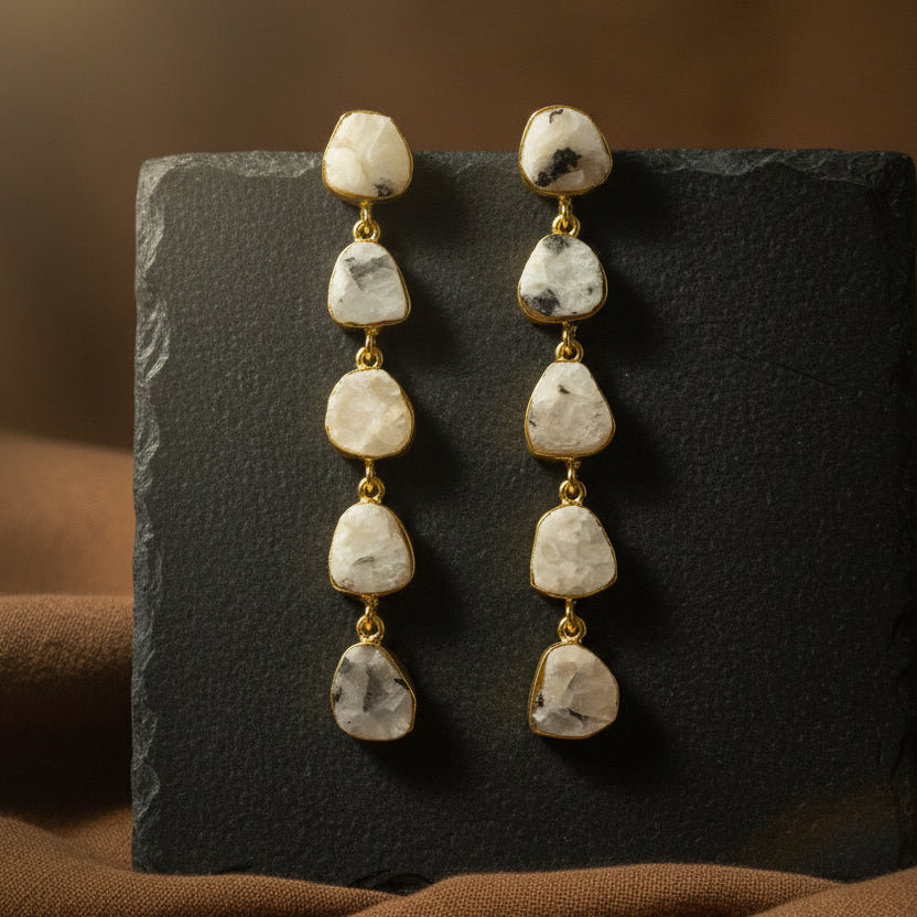 A pair of dangle earrings with irregular, polished gray stones in gold-toned settings, displayed on a textured black background.