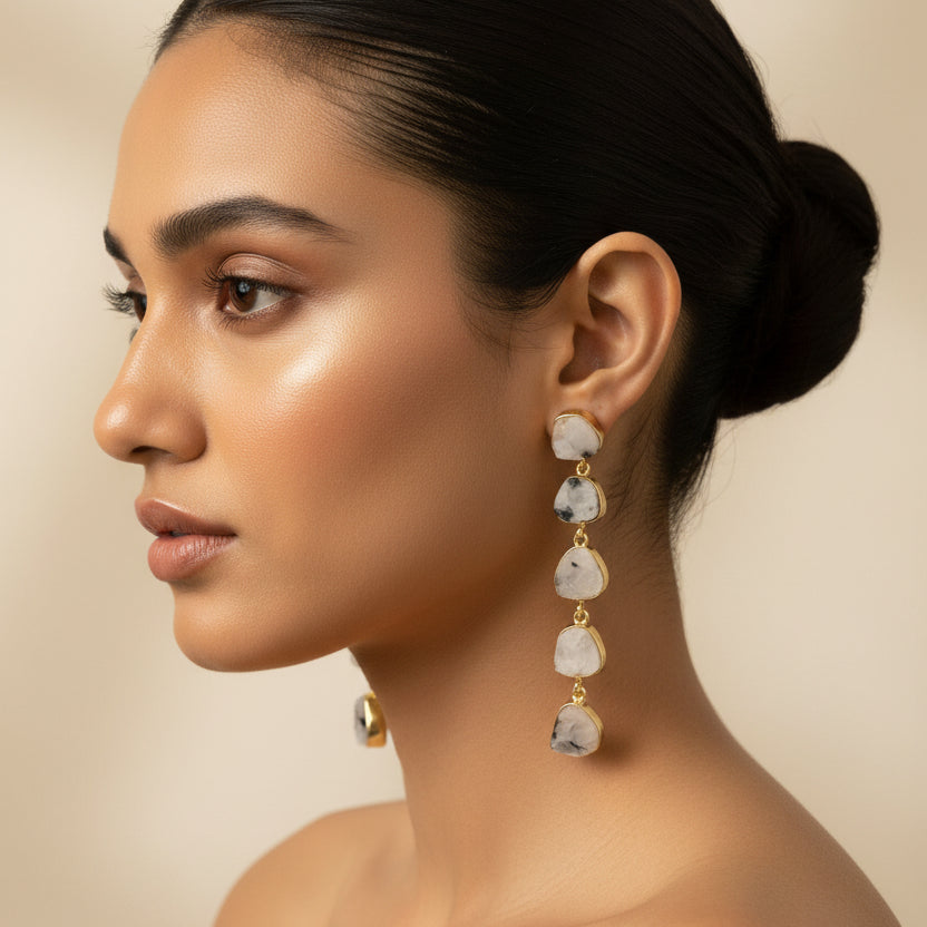A pair of dangle earrings with irregular, polished gray stones in gold-toned settings, displayed on a textured black background.