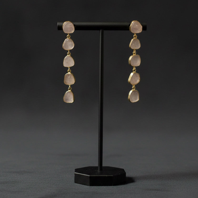 A pair of dangle earrings with irregular, translucent pale stone drops connected by small gold-toned beads, displayed on a textured black fabric background.