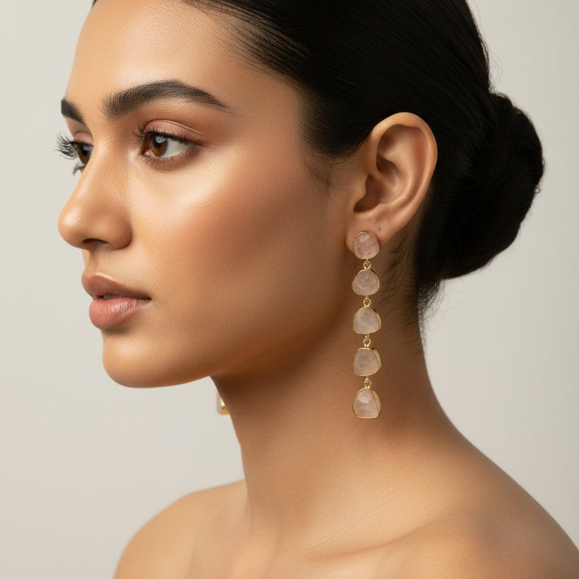 A pair of dangle earrings with irregular, translucent pale stone drops connected by small gold-toned beads, displayed on a textured black fabric background.