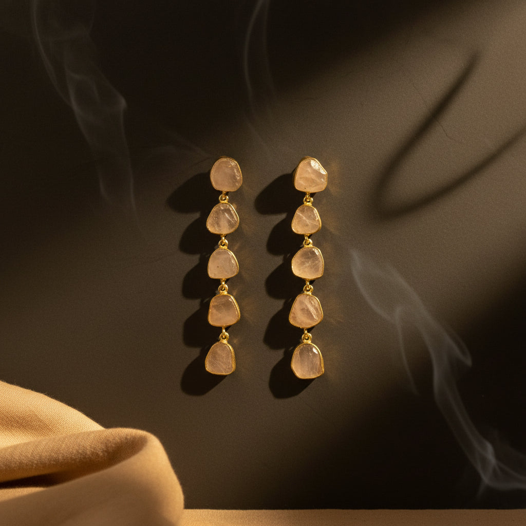 A pair of dangle earrings with irregular, translucent pale stone drops connected by small gold-toned beads, displayed on a textured black fabric background.