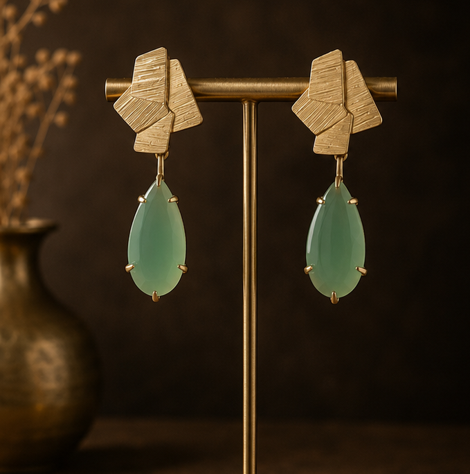 Geometric drop earrings with textured gold-tone metal and light green teardrop stones, shown on a display stand, worn by a model, and close-up on a dark stone surface.