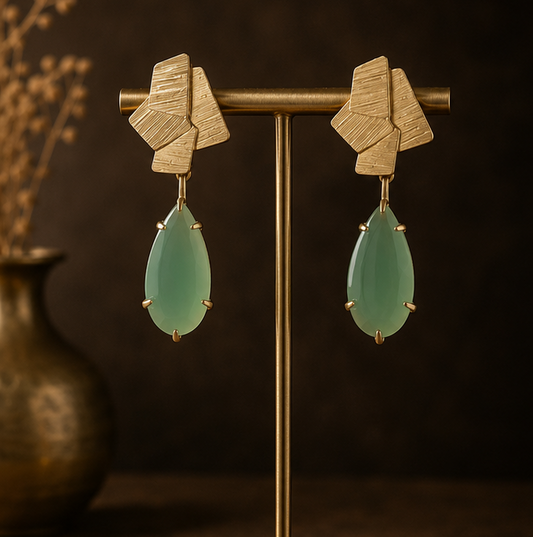 Geometric drop earrings with textured gold-tone metal and light green teardrop stones, shown on a display stand, worn by a model, and close-up on a dark stone surface.