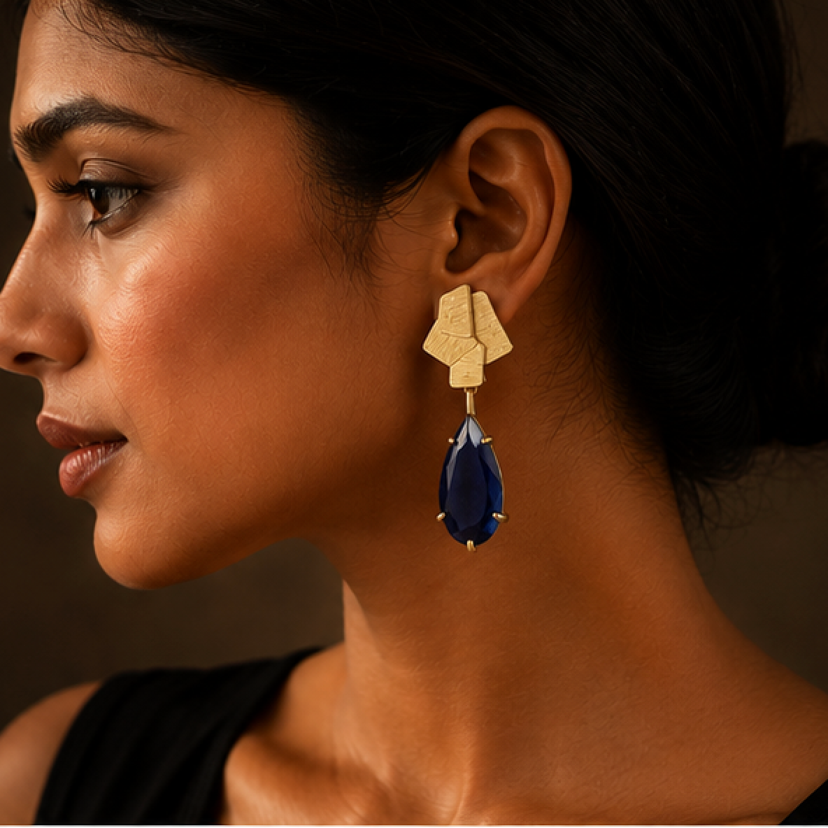 A close-up side profile of a woman wearing a statement earring with a textured gold geometric top and a deep teardrop-shaped dark blue drop, set against a dark brown background.