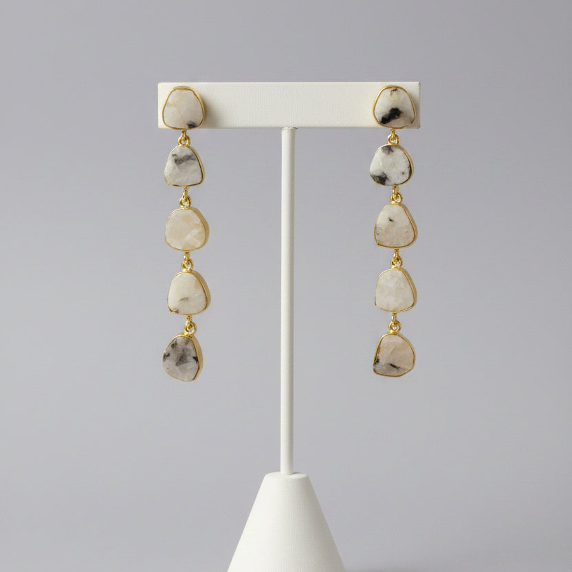 A pair of dangle earrings with irregular, polished gray stones in gold-toned settings, displayed on a textured black background.