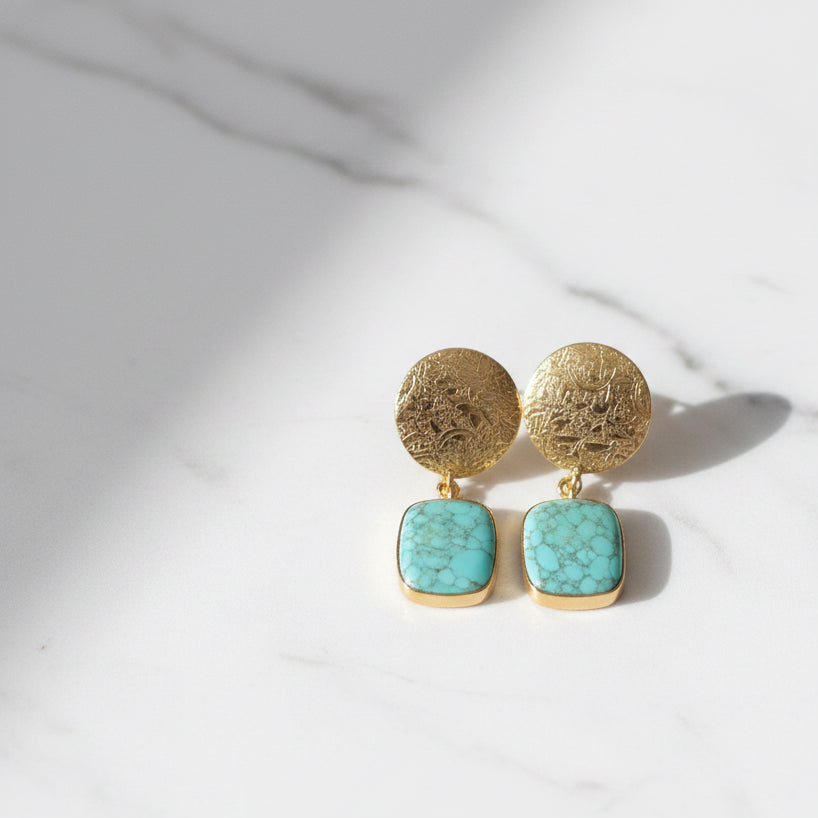 A pair of gold-toned dangle earrings with textured circular studs and square turquoise-blue stone drops, shown on a solid black background.