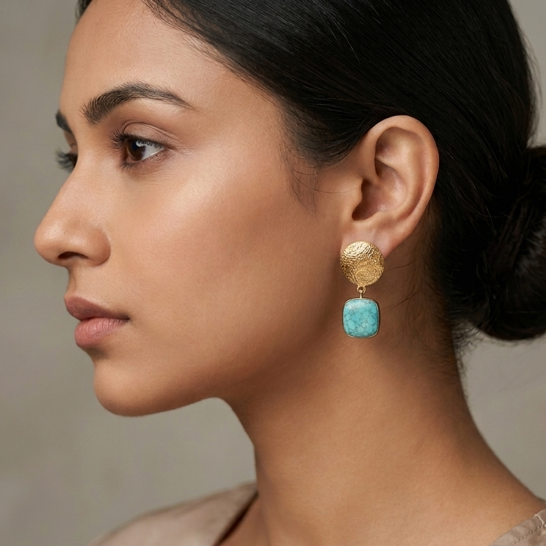 A woman in profile wearing a gold drop earring with a square turquoise blue stone, shown against a neutral gray background.