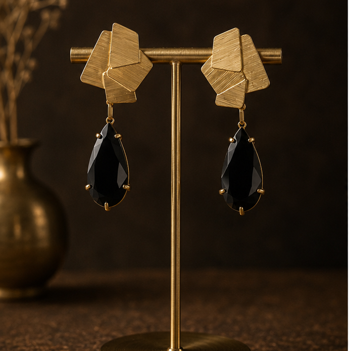 Geometric gold drop earrings with faceted gold top pieces and black teardrop-shaped drops, shown on a display stand, worn by a model, and on a dark textured surface.