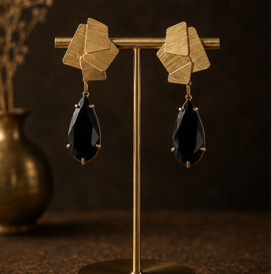 Geometric gold drop earrings with faceted gold top pieces and black teardrop-shaped drops, shown on a display stand, worn by a model, and on a dark textured surface.