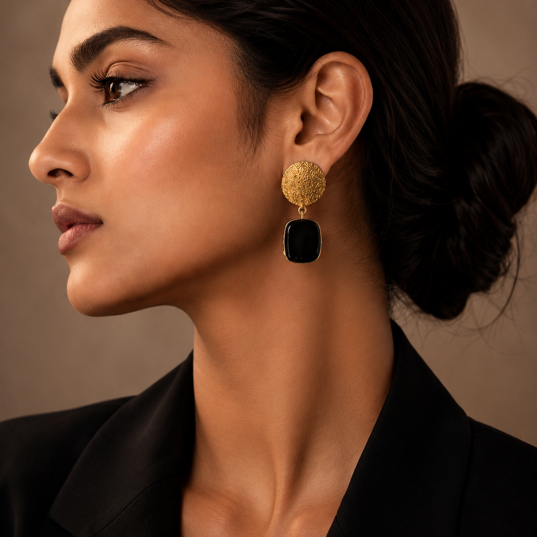 A close-up side profile of a woman wearing a gold and black drop earring, dressed in a black blazer against a warm brown background.