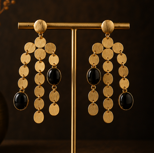 Gold-toned statement dangle earrings with circular coin-like discs and dark oval stones, shown on a display stand, worn by a model, and laid flat on a dark surface.