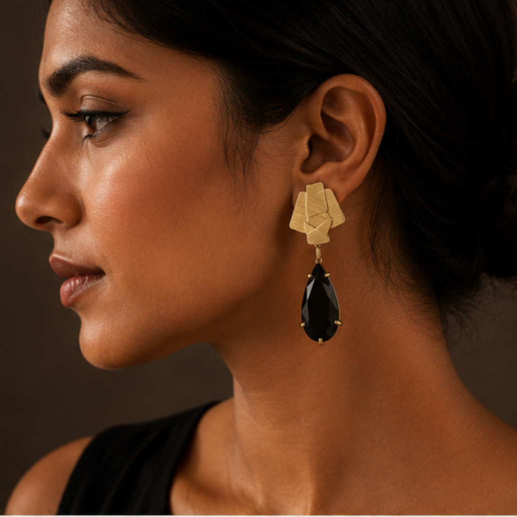 A close-up side profile of a woman wearing a geometric gold drop earring with a faceted black teardrop gemstone, set against a dark brown background.