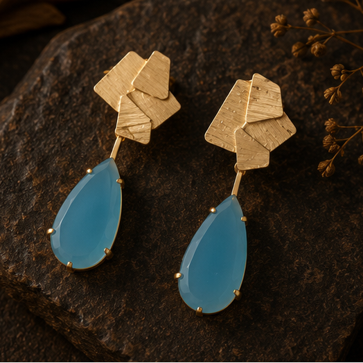 Geometric gold drop earrings with light blue teardrop-shaped stones, shown on a gold display stand, on a dark rock, and worn by a woman in a side-profile portrait.