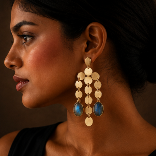 A close-up side profile of a woman wearing a large gold-toned dangle earring with circular disc charms and two blue oval stone drops, against a dark brown background.