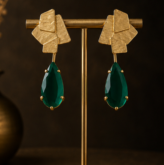 Geometric gold drop earrings with teardrop-shaped emerald green stones, shown on a gold display stand, worn by a model, and resting on a dark stone surface.