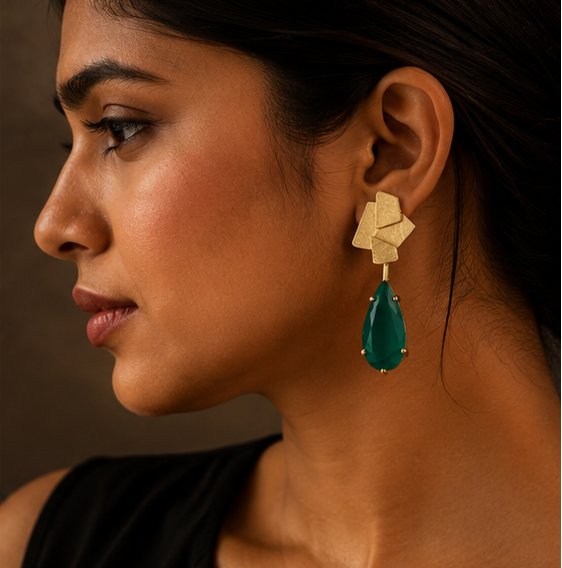 Geometric gold drop earrings with teardrop-shaped emerald green stones, shown on a gold display stand, worn by a model, and resting on a dark stone surface.