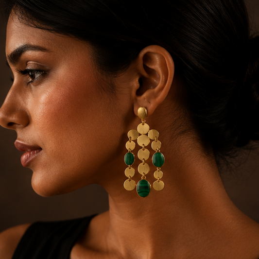 Gold-toned statement dangle earrings with oval gold disc drops and green stone accents, shown on a display stand and worn by a model in a dark warm-toned setting.