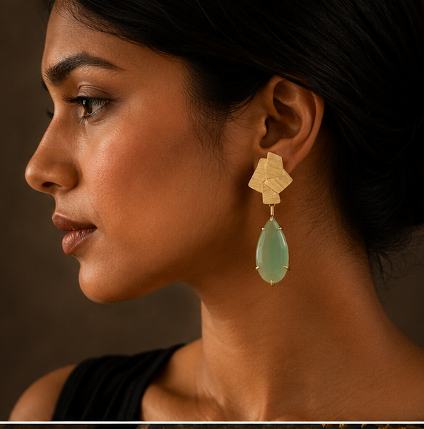 Geometric drop earrings with textured gold-tone metal and light green teardrop stones, shown on a display stand, worn by a model, and close-up on a dark stone surface.
