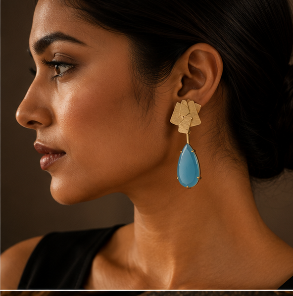 Geometric gold drop earrings with light blue teardrop-shaped stones, shown on a gold display stand, on a dark rock, and worn by a woman in a side-profile portrait.
