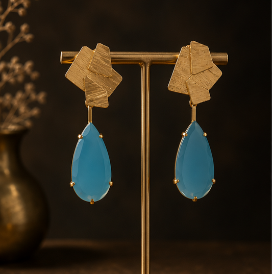 Geometric gold drop earrings with light blue teardrop-shaped stones, shown on a gold display stand, on a dark rock, and worn by a woman in a side-profile portrait.
