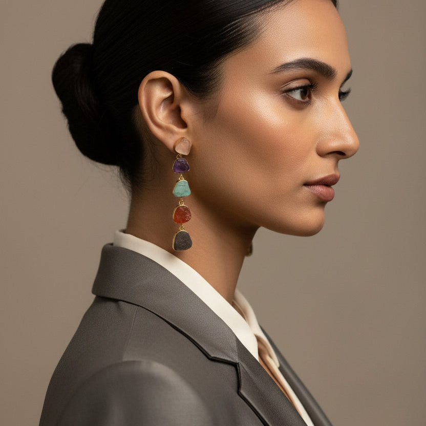 A pair of gold drop earrings featuring five small, irregular natural gemstones in purple, green, orange, gray, and beige, displayed on a black textured fabric background.