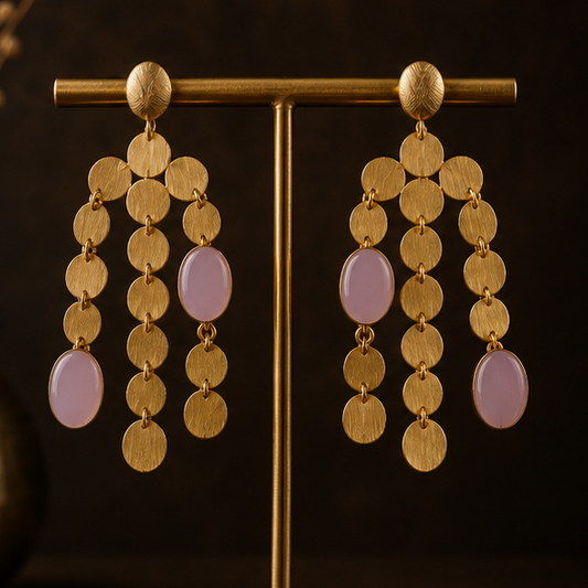 Gold-tone dangle earrings with layered circular coin-like discs and light pink oval drops, shown on a gold display stand, worn by a model in a side profile, and close-up on a dark textured surface.