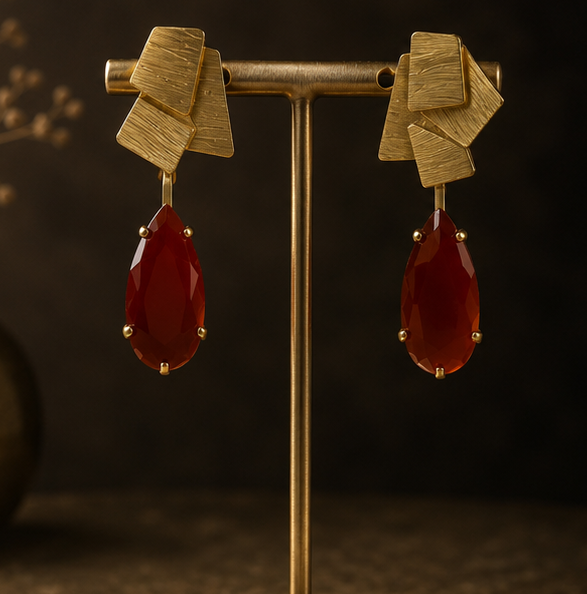 Collage showing a pair of statement earrings displayed on a gold stand, worn by a woman in a side-profile portrait, and resting on a dark textured surface. The earrings feature a textured gold geometric top with dangling teardrop-shaped deep red gemstones.