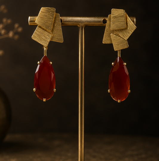 Collage showing a pair of statement earrings displayed on a gold stand, worn by a woman in a side-profile portrait, and resting on a dark textured surface. The earrings feature a textured gold geometric top with dangling teardrop-shaped deep red gemstones.