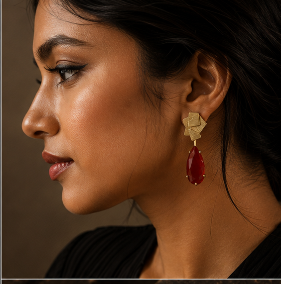 Collage showing a pair of statement earrings displayed on a gold stand, worn by a woman in a side-profile portrait, and resting on a dark textured surface. The earrings feature a textured gold geometric top with dangling teardrop-shaped deep red gemstones.