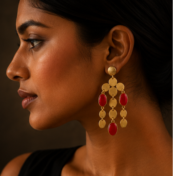 Gold-toned statement earrings with dangling oval and round gold discs and red teardrop beads, shown on a display stand, worn by a model in profile, and laid flat on a dark textured surface.